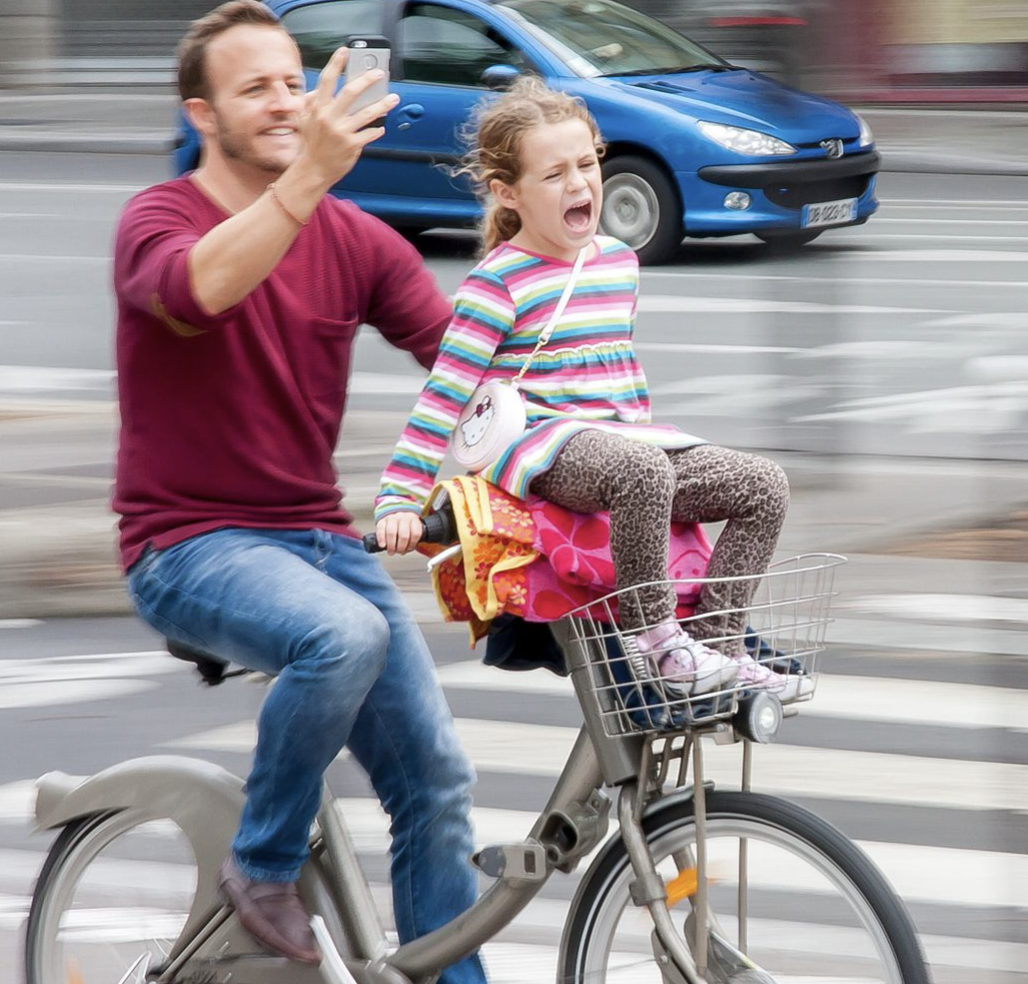 Man takes selfie while riding bicycle with child on handlebars