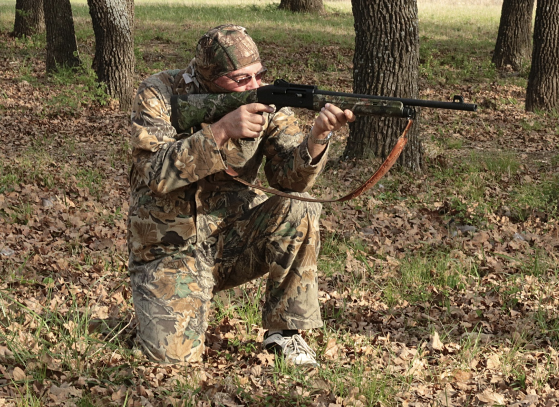 A man dressed in camouflage is aiming his shotgun as he is hunting in the woods.